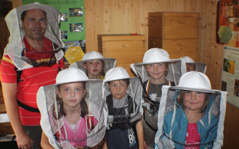 Eine Gruppe von Kindern und ein Erwachsener, die Imkeranzüge und Hüte tragen, stehen in einem Innenraum vor Holzkisten und Lehrplakaten.