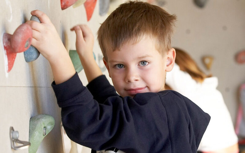 Ein kleiner Junge klettert in einer Halle an einer Felswand und schaut in die Kamera. Deutscher Text wirbt für Kinder- und Jugendferienkletterwochen in einer Salzburger Kletterhalle.