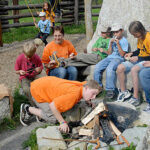 Eine Gruppe von Kindern und ein Erwachsener sitzen um eine Feuerstelle im Freien; ein Kind bläst auf das Feuer, während die anderen Stöcke halten.