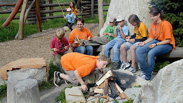Eine Gruppe von Kindern und ein Erwachsener sitzen um eine Feuerstelle im Freien; ein Kind bläst auf das Feuer, während die anderen Stöcke halten.