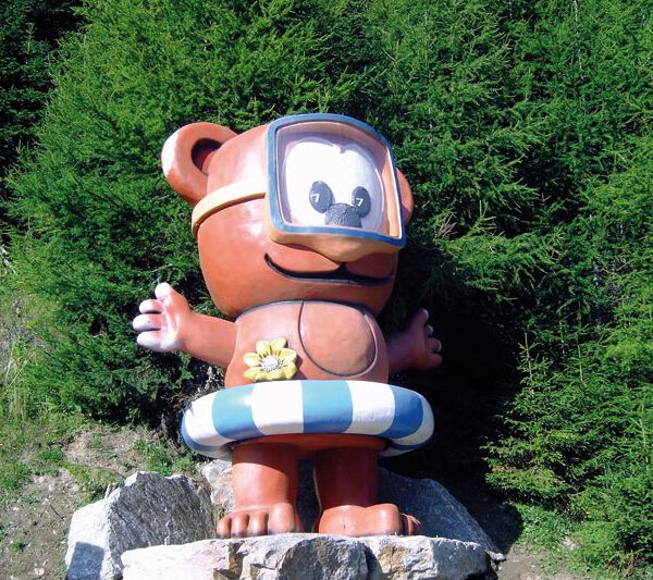 Eine große Cartoon-Bärenstatue mit Schwimmbrille und gestreiftem Schwimmring steht auf Felsen vor dichten grünen Bäumen.