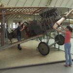 Zwei Personen interagieren mit einem ausgestellten Doppeldecker aus der Zeit des Ersten Weltkriegs; eine Person dreht den Propeller, während die andere das Cockpit untersucht. Der Rahmen und der Motor des Flugzeugs sind ausgestellt.