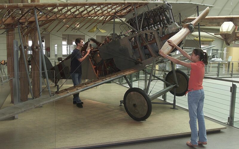 Zwei Personen interagieren mit einem ausgestellten Doppeldecker aus der Zeit des Ersten Weltkriegs; eine Person dreht den Propeller, während die andere das Cockpit untersucht. Der Rahmen und der Motor des Flugzeugs sind ausgestellt.