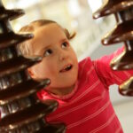 Ein junges Mädchen in einem rosafarbenen Hemd blickt mit einem neugierigen Gesichtsausdruck zwischen zwei großen braunen Isolatoren hervor.