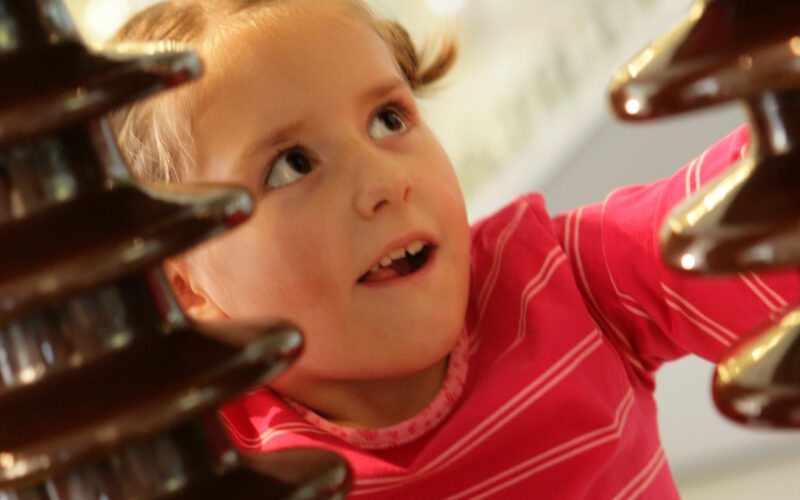 Ein junges Mädchen in einem rosafarbenen Hemd blickt mit einem neugierigen Gesichtsausdruck zwischen zwei großen braunen Isolatoren hervor.