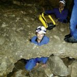 Eine Person in Höhlenausrüstung hockt durch einen engen Felsdurchgang in einer Höhle, während eine andere Person in der Nähe steht, beide tragen Helme mit Stirnlampen.