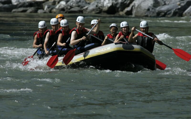 Eine Gruppe von Personen mit Helmen und Schwimmwesten paddelt auf einem Schlauchboot durch einen Fluss mit felsigen Ufern.