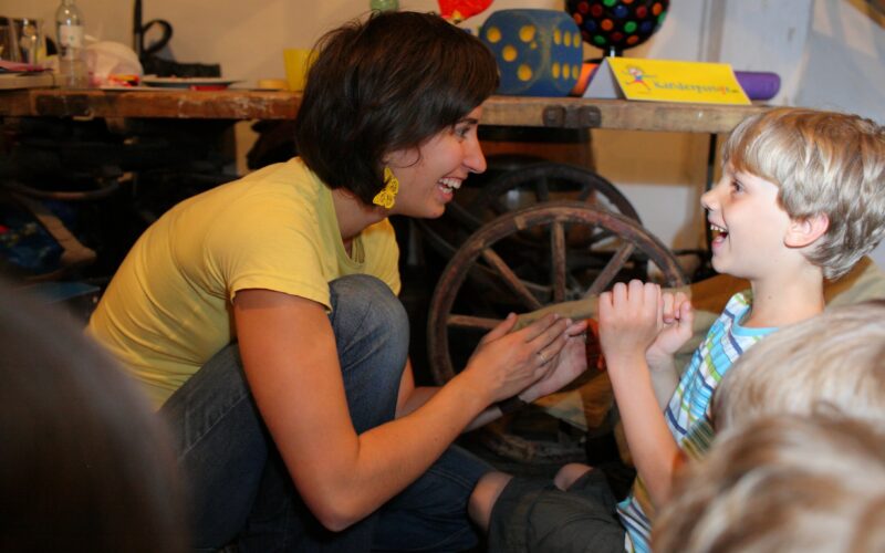 Eine Frau und ein kleiner Junge sitzen lächelnd auf dem Boden im Haus. Im Hintergrund sind ein hölzernes Wagenrad und bunte Gegenstände zu sehen.
