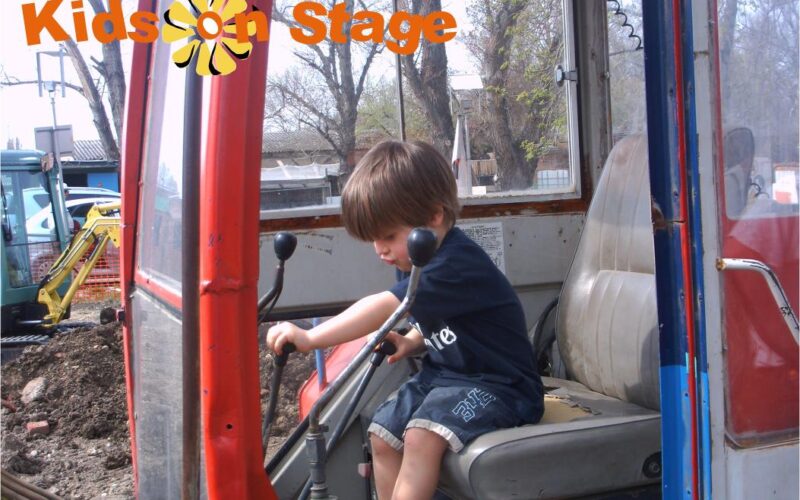 Ein kleines Kind sitzt auf dem Fahrersitz einer Baumaschine und hält zwei Steuerhebel in der Hand. Der Schriftzug "Kids on Stage" erscheint oben links. Rechts unten steht das Datum "2009-04-05".