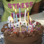 Ein Schokoladengeburtstagskuchen mit brennenden Kerzen und Süßigkeiten steht auf einem Tisch; im Hintergrund ist eine Person in einem gelben Hemd zu sehen.