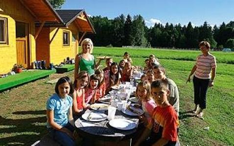 Eine Gruppe von Kindern und zwei Erwachsene sitzen an einem langen Tisch im Freien, der für eine Mahlzeit in der Nähe eines Holzgebäudes an einem sonnigen Tag gedeckt ist.