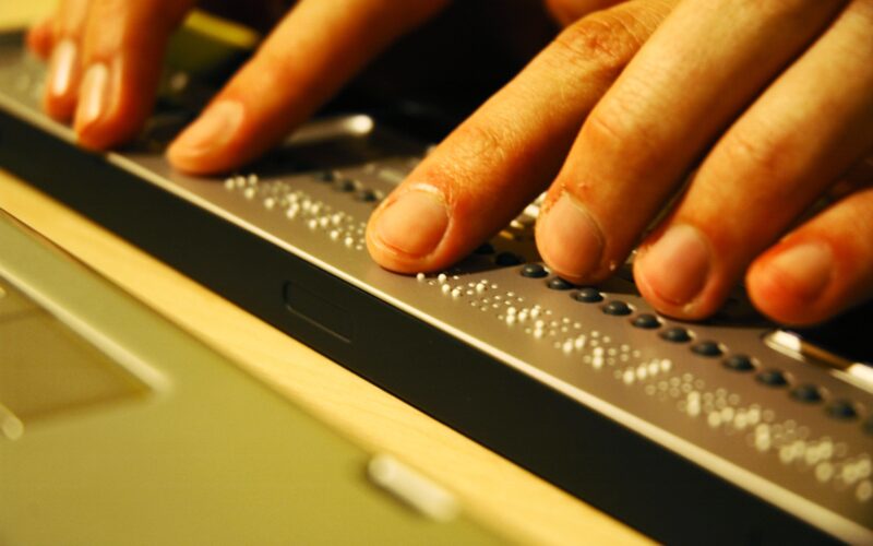 Nahaufnahme von Händen, die Blindenschrift auf einer aktualisierbaren Braillezeile neben einer Laptop-Tastatur lesen.