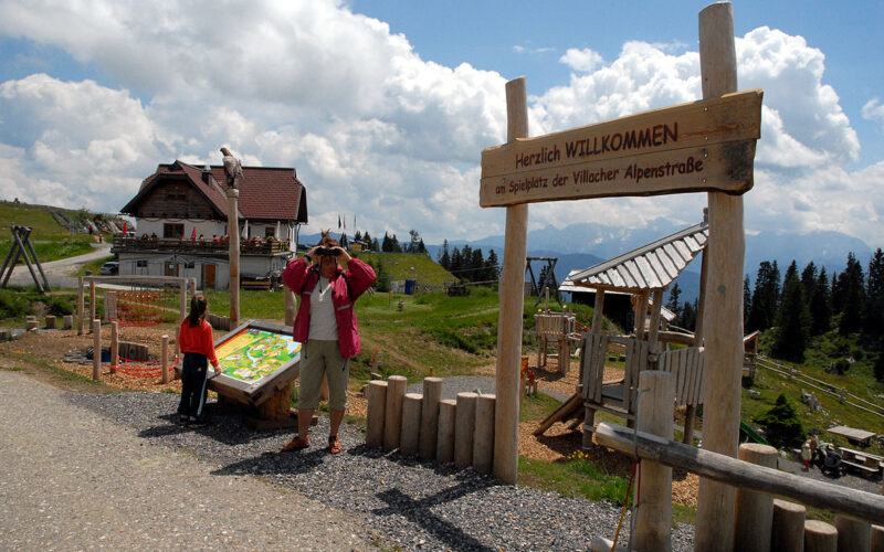Ein Mann macht ein Foto in der Nähe eines hölzernen Willkommensschildes an einem Aussichtspunkt in den Bergen, mit einem Kind und einem Gebäude im Chalet-Stil im Hintergrund unter einem teilweise bewölkten Himmel.