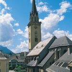 Eine hohe Kirche mit einem spitzen Kirchturm steht im Zentrum einer kleinen Stadt unter einem strahlend blauen Himmel mit vereinzelten Wolken.