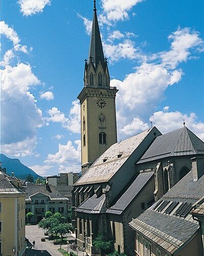 Eine hohe Kirche mit einem spitzen Kirchturm steht im Zentrum einer kleinen Stadt unter einem strahlend blauen Himmel mit vereinzelten Wolken.
