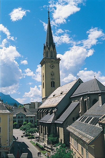 Eine hohe Kirche mit einem spitzen Kirchturm steht im Zentrum einer kleinen Stadt unter einem strahlend blauen Himmel mit vereinzelten Wolken.