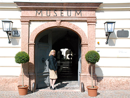 Eine Frau steht am Eingang eines Museums mit einem gewölbten Portal, flankiert von zwei Topfpflanzen und zwei an der Wand befestigten Lampen.