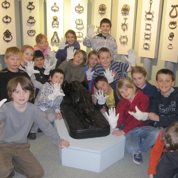 Eine Gruppe von Kindern mit weißen Handschuhen posiert mit einer schwarzen Statue in einem Museum, hinter der dekorative Gegenstände an den Wänden ausgestellt sind.