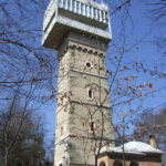 Ein hoher, rechteckiger Steinturm mit einer großen weißen Struktur auf der Spitze, umgeben von blattlosen Bäumen und einem geparkten Auto, unter einem klaren blauen Himmel.