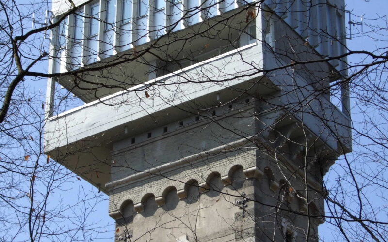 Ein hoher Steinturm mit einer modernen gläsernen Aussichtsplattform auf der Spitze, die man durch kahle Äste vor einem klaren blauen Himmel sieht.