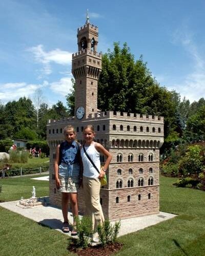 Zwei Mädchen stehen an einem sonnigen Tag vor einem detaillierten Miniaturmodell eines schlossähnlichen Gebäudes im Freien.