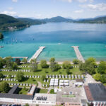 Luftaufnahme eines Seeufers mit klarem, blauem Wasser, mehreren Anlegestellen, grünen Parkanlagen, kleinen Gebäuden und umliegenden Hügeln unter einem teilweise bewölkten Himmel.