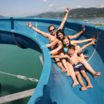 Vier Personen in Badeanzügen fahren gemeinsam eine blaue Wasserrutsche über ein großes Gewässer hinunter, mit Bergen und einem teilweise bewölkten Himmel im Hintergrund.
