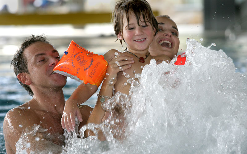 Drei Personen, darunter ein Kind mit orangefarbenen Schwimmflügeln, lächeln und spielen zusammen in einem Schwimmbad, während das Wasser um sie herum aufspritzt.