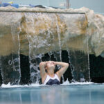 Eine Person im Badeanzug steht unter einem steinernen Wasserfall in einem Schwimmbecken, die Hände auf dem Kopf und die Augen geschlossen.
