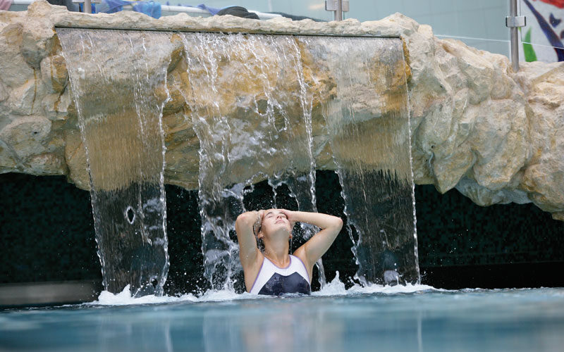 Eine Person im Badeanzug steht unter einem steinernen Wasserfall in einem Schwimmbecken, die Hände auf dem Kopf und die Augen geschlossen.