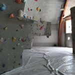 Indoor-Kletterhalle mit strukturierten Kletterwänden und bunten Griffen, gepolstertem Boden, rotem Vorhang und großen Fenstern, die viel Tageslicht hereinlassen.