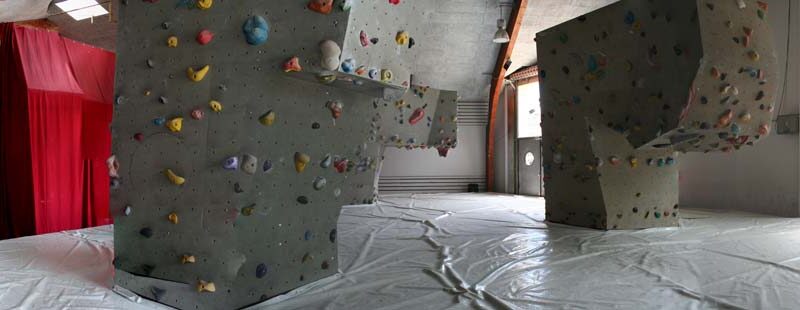Indoor-Kletterhalle mit strukturierten Kletterwänden und bunten Griffen, gepolstertem Boden, rotem Vorhang und großen Fenstern, die viel Tageslicht hereinlassen.