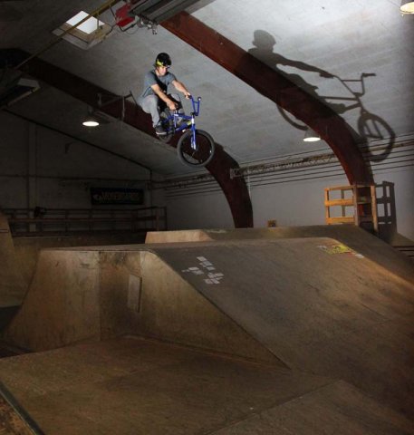 Eine Person fährt mit einem BMX-Rad von einer Rampe in einem Indoor-Skatepark und wirft dabei einen Schatten auf die gewölbte Wand hinter ihr.