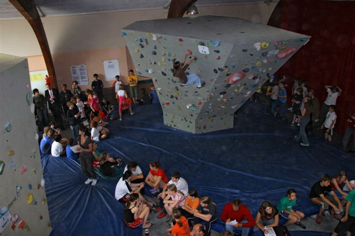 Boulder-Wettbewerb in der Halle mit Kletterern an einer Kletterwand und einer Menge von Zuschauern, die auf blauen Matten sitzen und stehen.