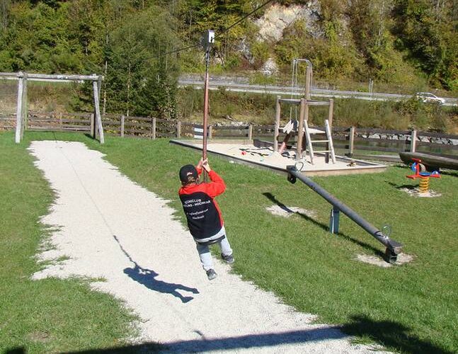Ein Kind in einer rot-schwarzen Jacke fährt mit einer Seilrutsche auf einem Spielplatz mit Gras, Kieswegen und Spielgeräten.