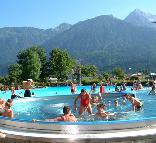 Menschen verschiedenen Alters schwimmen und spielen in einem Freibad, umgeben von Grün und Bergen unter einem klaren Himmel.