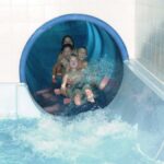 Vier Kinder kommen aus einer blauen, geschlossenen Wasserrutsche in ein Schwimmbecken und spritzen Wasser, während sie die Rutsche verlassen.