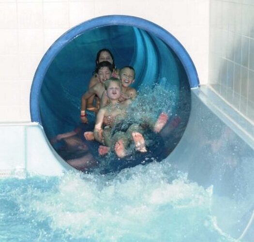Vier Kinder kommen aus einer blauen, geschlossenen Wasserrutsche in ein Schwimmbecken und spritzen Wasser, während sie die Rutsche verlassen.