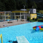 Freibad mit einem bunten aufblasbaren Spielzeug, einem gelben Sprungbrett und einem modernen Gebäude mit blauen Wasserrutschen im Hintergrund, umgeben von Bäumen und Grünanlagen.