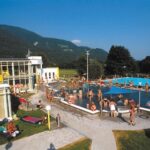 Öffentliches Freibad mit vielen Menschen, die schwimmen, sich sonnen und entspannen; umgeben von Gras, Bäumen und Bergen unter einem klaren blauen Himmel.