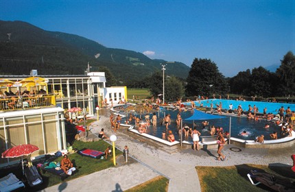 Öffentliches Freibad mit vielen Menschen, die schwimmen, sich sonnen und entspannen; umgeben von Gras, Bäumen und Bergen unter einem klaren blauen Himmel.