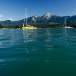 Zwei Segelboote treiben auf einem ruhigen See mit klarem Wasser, umgeben von grünen Hügeln und Bergen unter einem strahlend blauen Himmel.