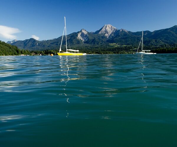 Zwei Segelboote treiben auf einem ruhigen See mit klarem Wasser, umgeben von grünen Hügeln und Bergen unter einem strahlend blauen Himmel.
