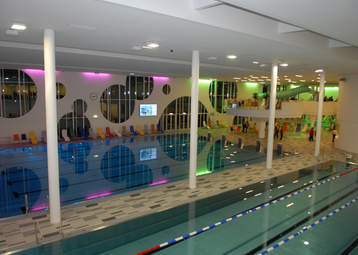 Hallenbad mit bunten Liegestühlen, modernem Dekor und farbigen Lichtern, die sich auf dem Wasser spiegeln.