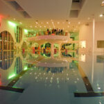 Hallenbad mit moderner Architektur, farbenfroher Beleuchtung, Liegestühlen und einer Gruppe von Personen, die auf einer erhöhten Aussichtsplattform stehen.