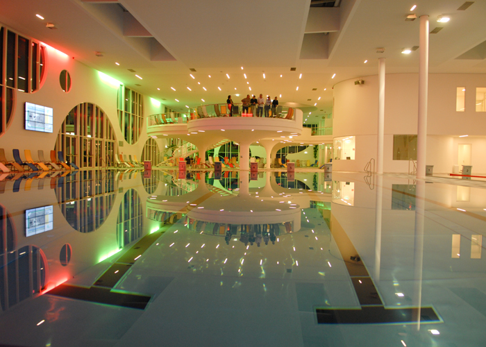 Hallenbad mit moderner Architektur, farbenfroher Beleuchtung, Liegestühlen und einer Gruppe von Personen, die auf einer erhöhten Aussichtsplattform stehen.