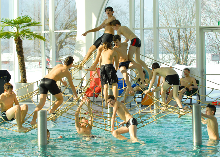 Eine Gruppe von Jungen in Badekleidung klettert und spielt auf einer Seilstruktur über einem Becken in einem Indoor-Badezentrum mit großen Fenstern und einer Palme.