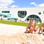 Drei Kinder in Badeanzügen knien an einem sonnigen Tag auf sandigem Boden vor einem modernen Gebäude mit großen Fenstern und Wasserrutschen im Freien.