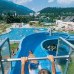 Zwei Kinder rutschen eine blaue Wasserrutsche hinunter in ein Freibad, umgeben von Menschen, Grasflächen und Bergen im Hintergrund.
