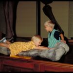 Zwei kleine Kinder interagieren mit einem großen Dinosaurierknochen, der auf einer hölzernen Oberfläche in einem Museum ausgestellt ist.